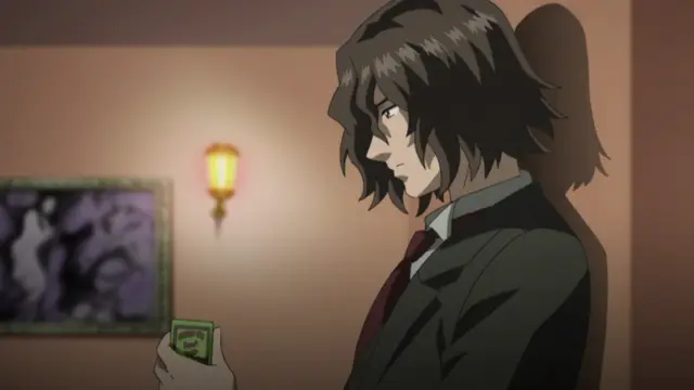 A medium shot features a dark-haired man in a suit holding a small green card with a face on it, standing in a dimly lit room with a picture on the wall. Screenshot from Kara no Shoujo - Episode 1