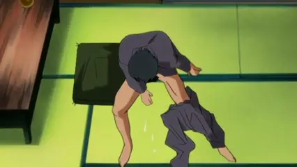 A muscular man with dark hair is bent over on a tatami mat floor in a traditional Japanese room, his pants around his ankles and fluid dripping from his crotch as he bows toward a dark green cushion. Screenshot from Hana Dorei - Episode 1