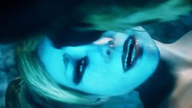 A close-up shot of a blonde woman with wide eyes and an open mouth, lying horizontally under a dark, translucent blue surface. Screenshot from Scarlet Nights