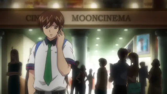 A brown-haired anime boy in a school uniform stands in front of a cinema called "MOONCINEMA" while talking on a cell phone, surrounded by several other people in a lobby. Screenshot from Inshitsu Otaku ni Ikareru Kanojo - Episode 2