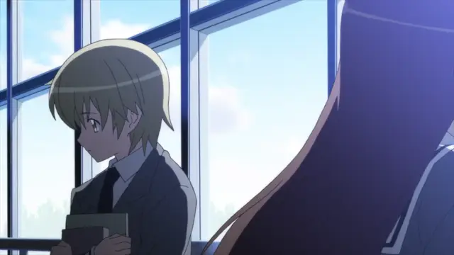 A blond male student in a school uniform looks down sadly while holding a book in a classroom, with the back of a long-haired girl's head visible in the foreground. Screenshot from Aki Sora: In a Dream - Episode 1