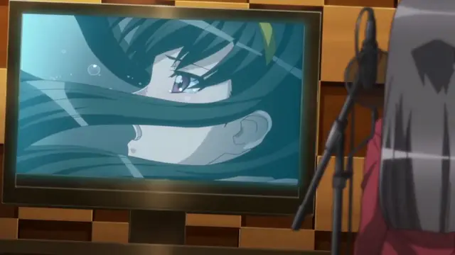 A young girl with long black hair and large eyes looks toward the viewer on a television screen in a room with wood-paneled walls, with a person's shoulder and a microphone visible in the foreground. Screenshot from Yume Kui: Tsurumiku Shiki Game Seisaku - Episode 1