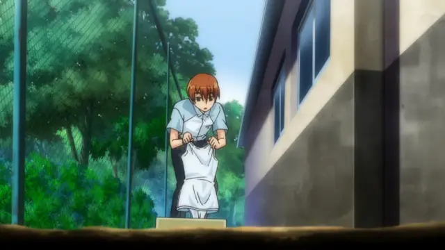 A young woman with short brown hair, wearing a white apron over a school uniform, sits on a stool outside a building next to a chain-link fence and trees. Screenshot from Tsundere Inran Shoujo Sukumi - Episode 1